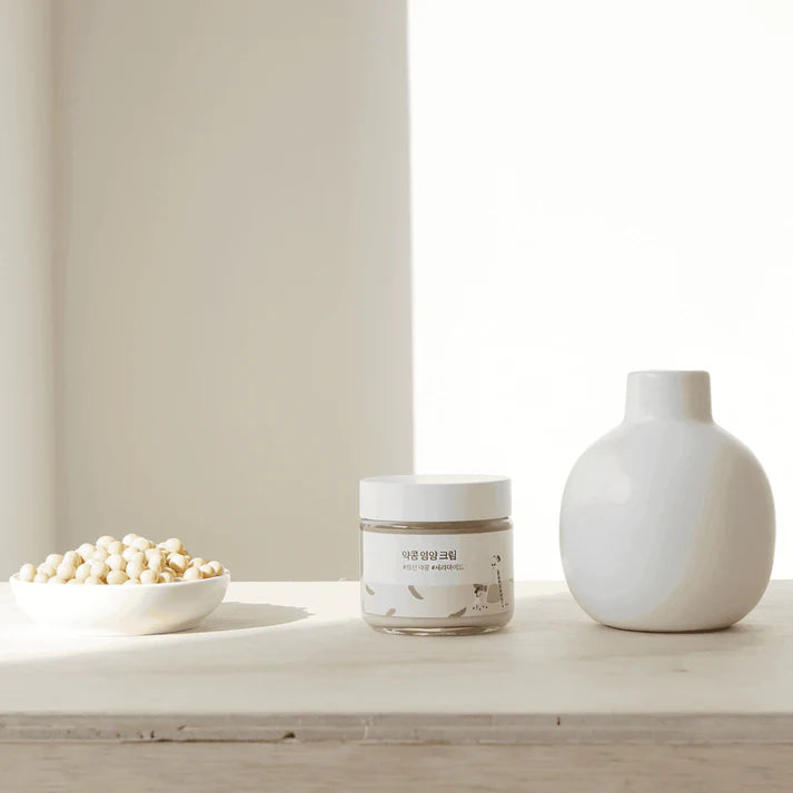 Jar of cream, bowl of nuts, and white vase on a light wooden surface with a neutral background