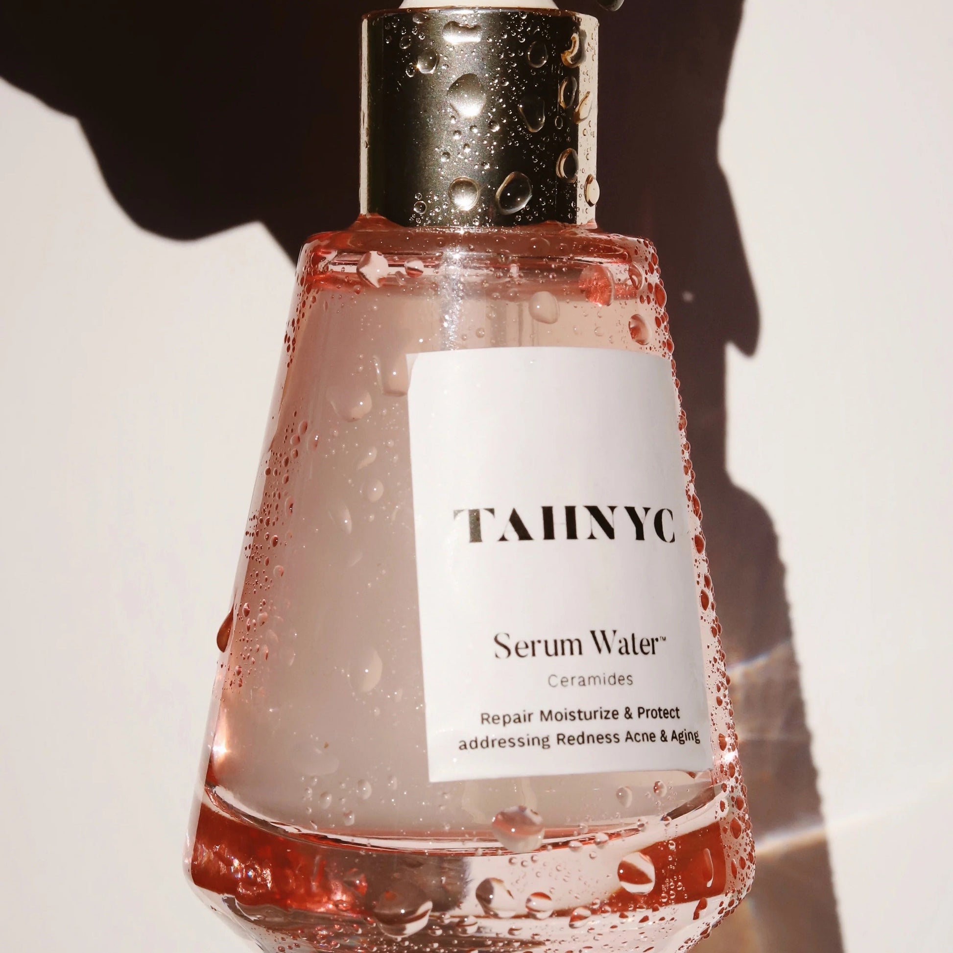 Bottle of TAHNYC Serum Water held by a hand with green nail polish on a neutral background