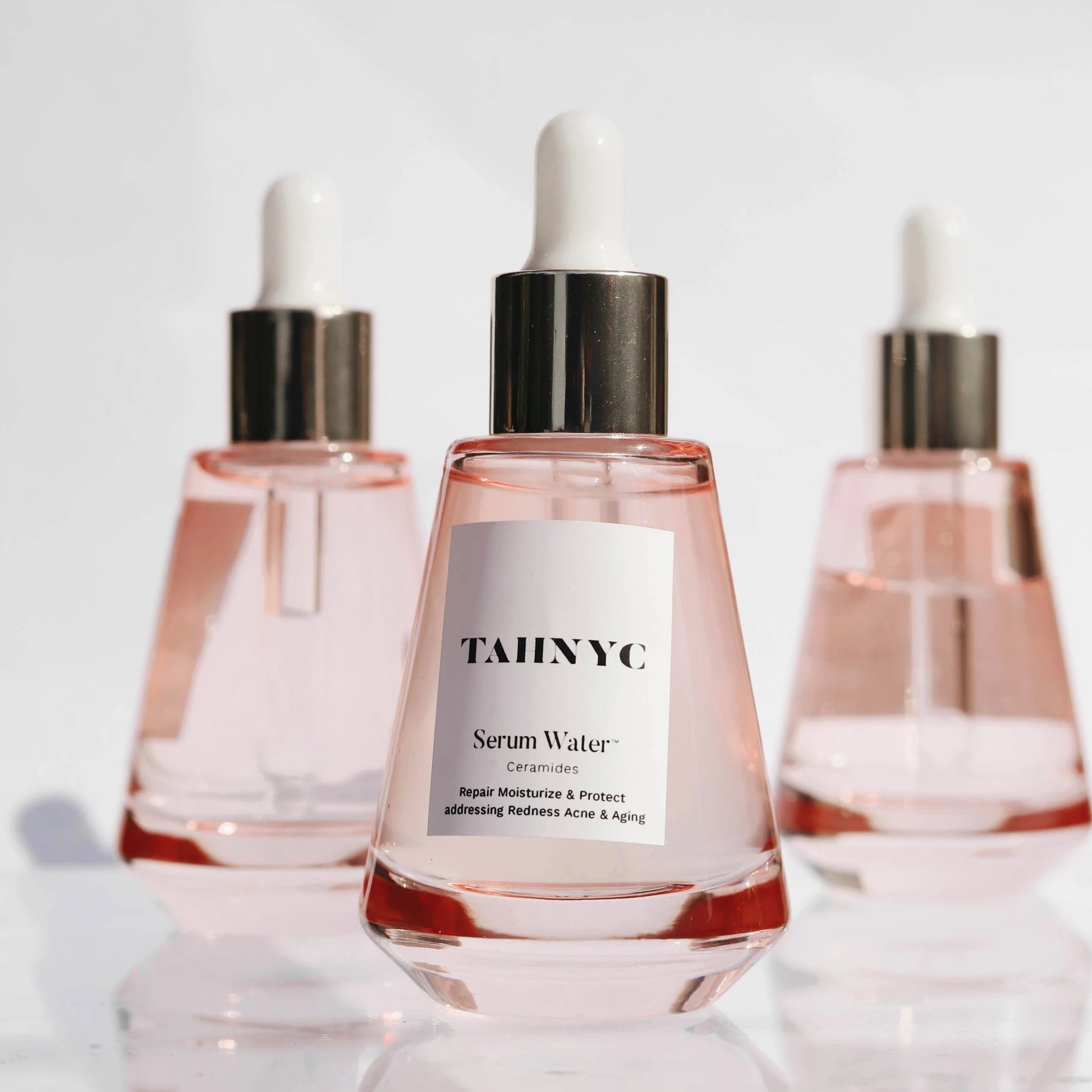 Three bottles of TAHNYC Serum Water on a reflective surface with a light gray background