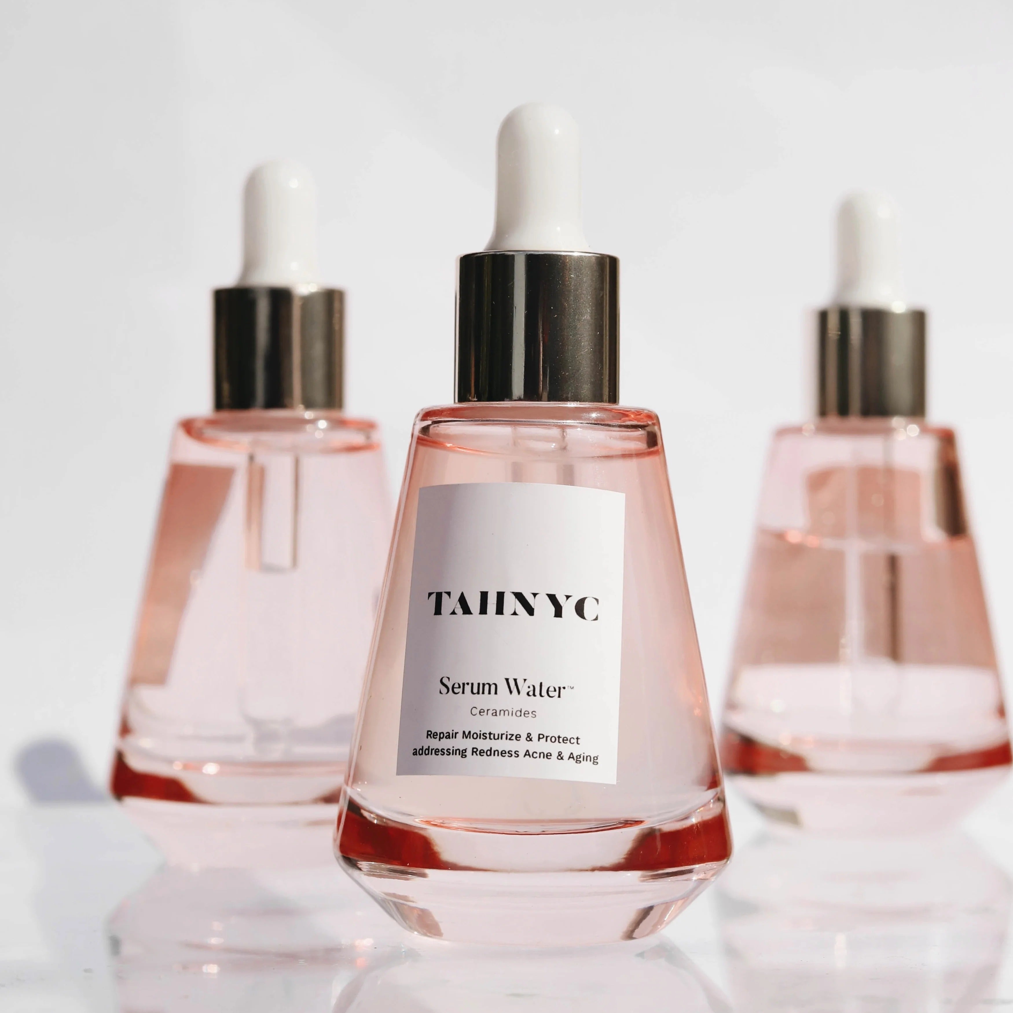 Three bottles of TAHNYC Serum Water on a reflective surface with a light gray background