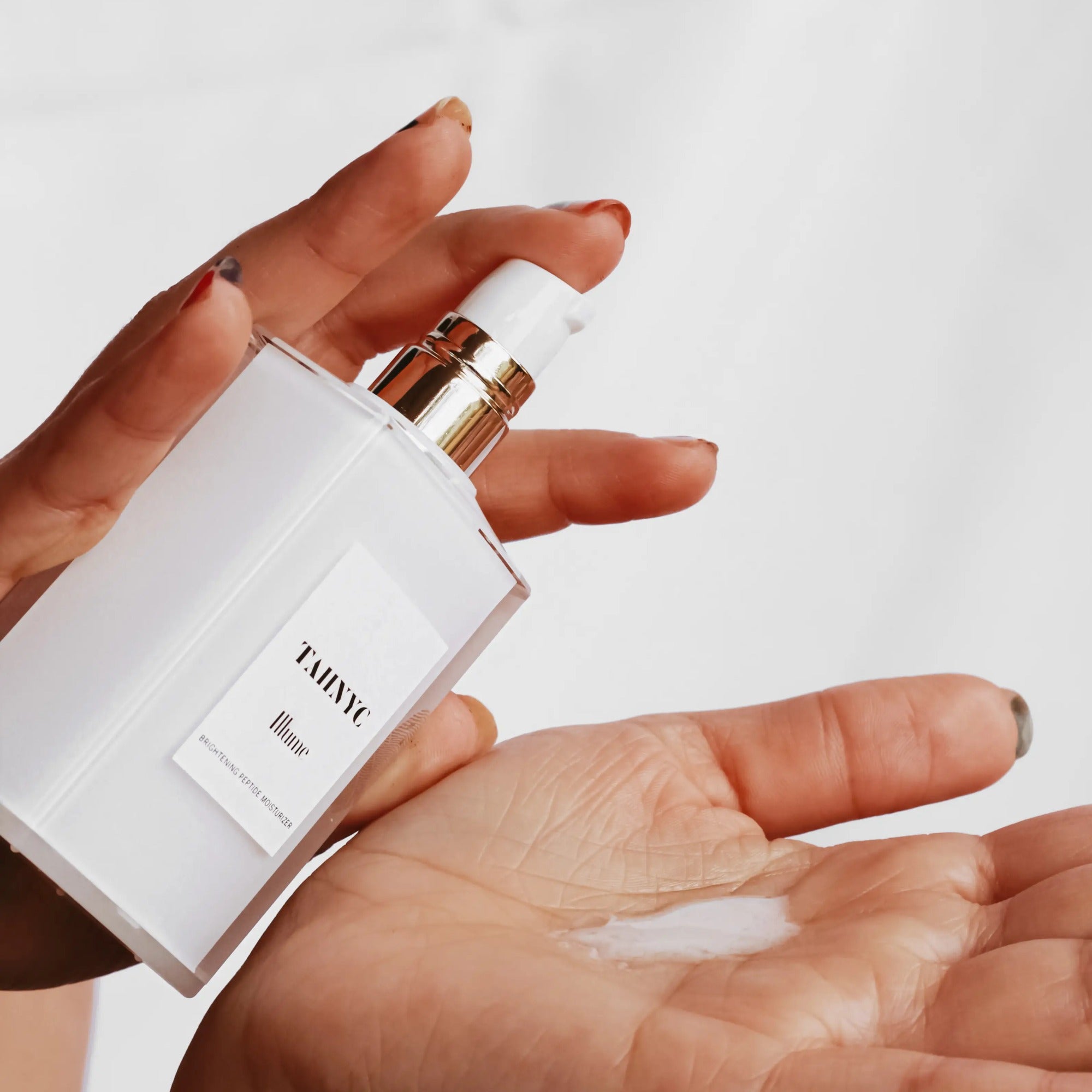 Hand holding a white skincare product with gold accents, showing cream on palm against a light background.