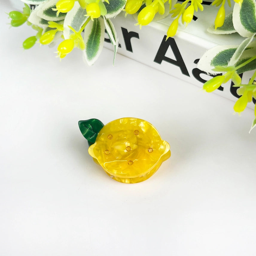 Yellow lemon-shaped object with a green leaf on a white surface with green leaves in the background.