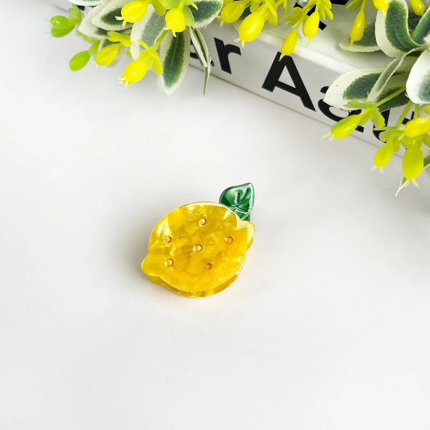 Yellow lemon-shaped earring with green leaf on a white surface with decorative plants.