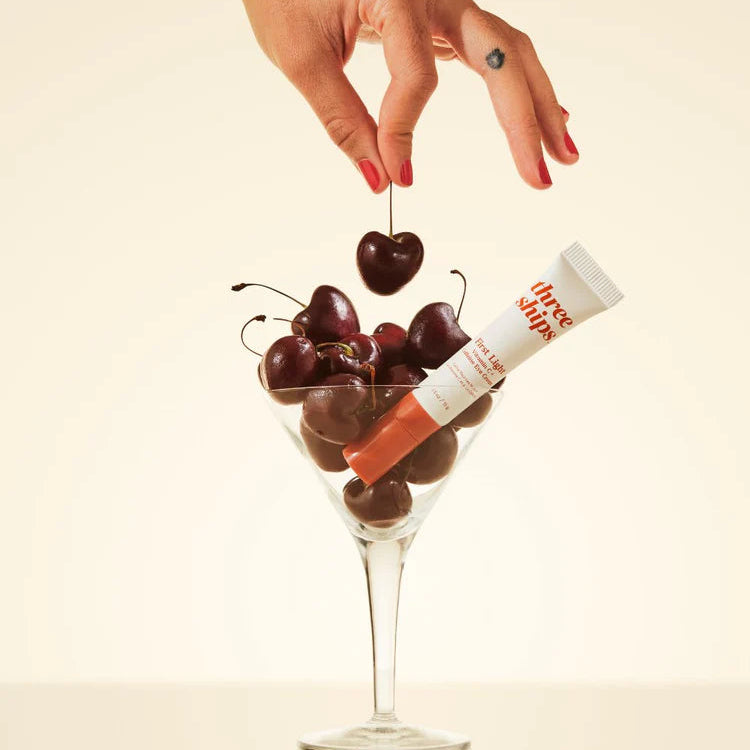 Hand holding a cherry above a glass filled with cherries and a tube of lip balm on a beige background