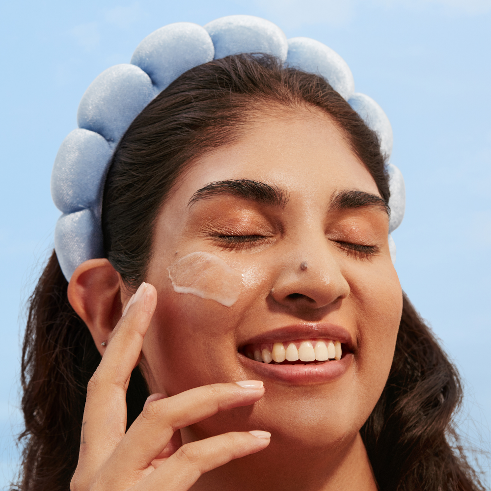 Woman applying cream to her face with a blue headband against a clear sky.