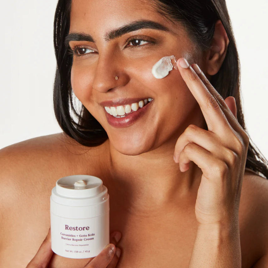 Woman applying cream to her face with a 'Restore' jar in hand against a neutral background