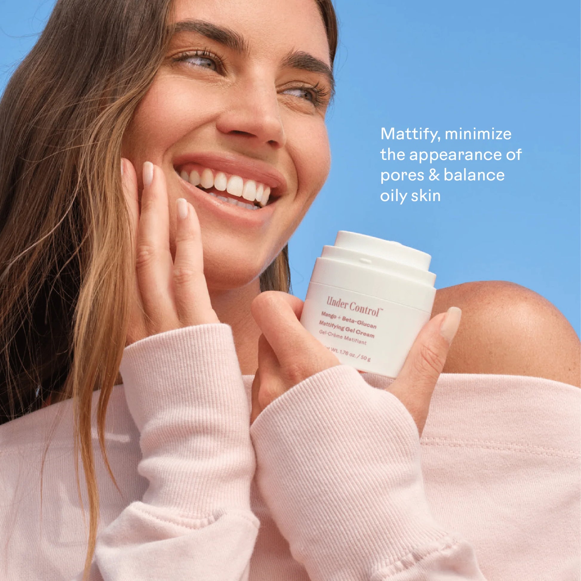 A smiling woman with long brown hair holds a white Under Control moisturizer jar against a bright blue sky. She wears a light pink sweatshirt and touches her face, with white text on the right reading, 'Mattify, minimize the appearance of pores & balance oily skin.