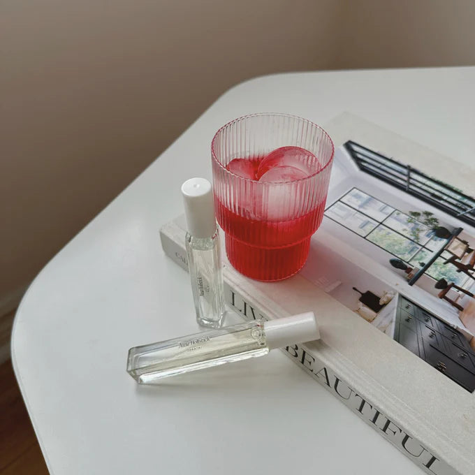 Red glass with ice cubes on a magazine next to a perfume bottle on a white surface.
