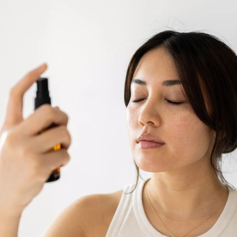 A woman with her eyes closed holds a small black spray bottle near her face, appearing to mist a fragrance or skincare product. She is wearing a white tank top against a clean white background.