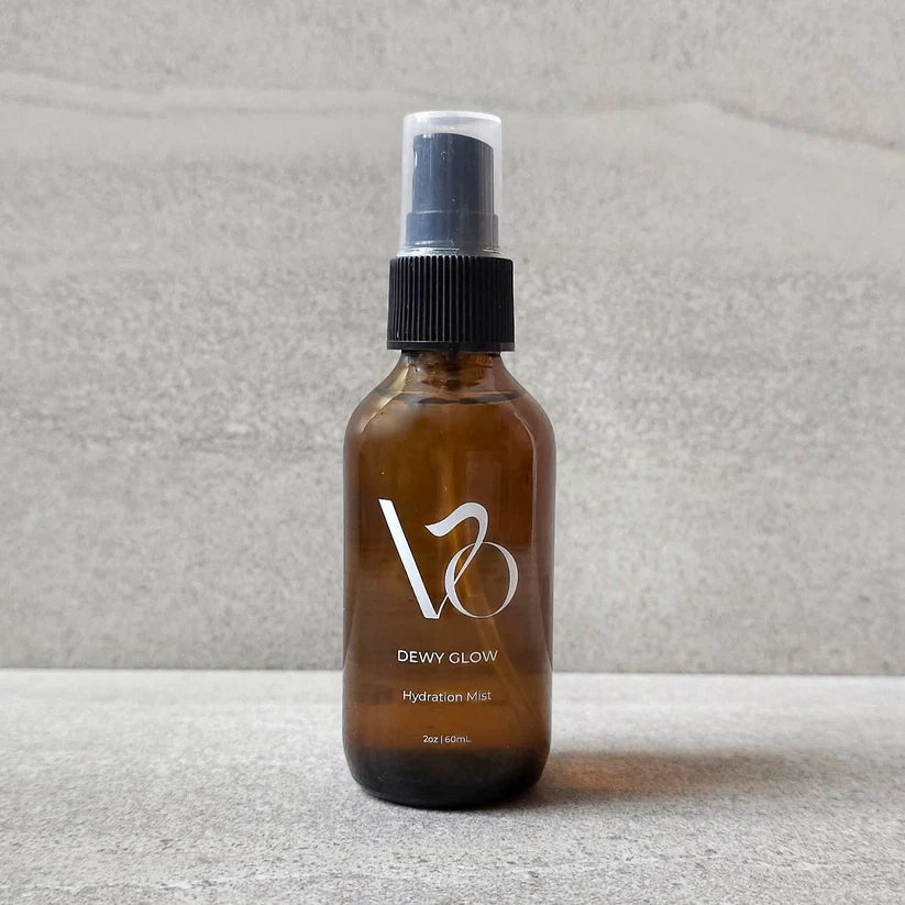 An amber glass spray bottle with a black pump and clear cap, labeled 'V8 DEWY GLOW Hydration Mist', standing on a gray textured surface against a matching gray background.