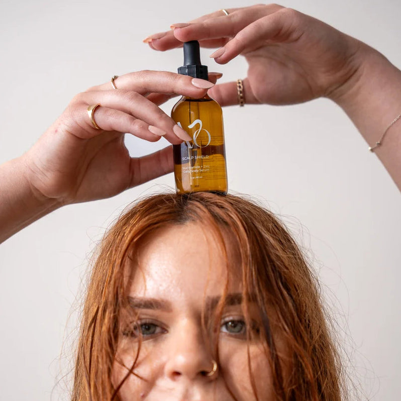 A woman with wet, reddish-brown hair holds a small amber dropper bottle above her forehead against a plain light background.